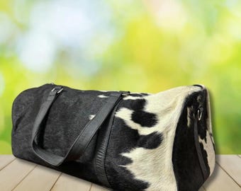 May include: A black and white cowhide duffel bag with black leather handles and trim. The bag is cylindrical and rests on a wooden surface. The background is a blurred mix of green and yellow.
