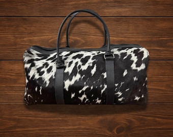 May include: A black and white cowhide leather duffel bag with black leather handles and straps.