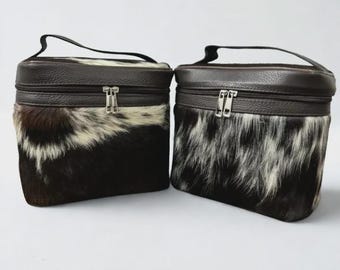 May include: Two matching cosmetic bags. The bags have a dark brown leather top and handle, with a zipper closure. The lower portion of the bags is made of black and white cowhide. The bags are rectangular with rounded corners.