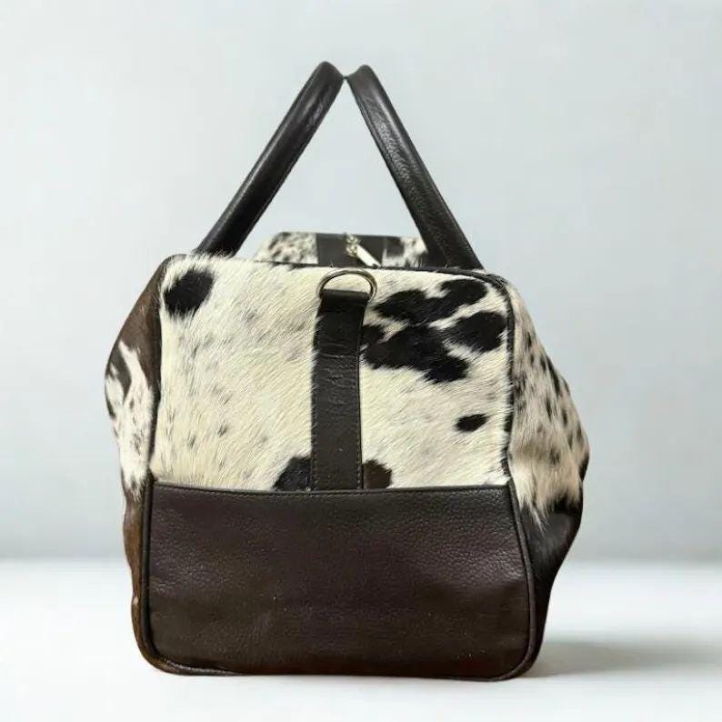 May include: A rectangular duffel bag with a cowhide pattern in black and white, and dark brown leather accents. The bag has two top handles and a dark brown leather base. A dark brown leather strap with a metal ring is attached to the front.
