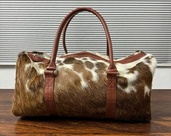 May include: A brown and white cowhide duffel bag with brown leather handles and straps. The bag has a zipper closure and is designed for travel or carrying items. The bag is sitting on a wooden surface.