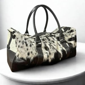 May include: A duffel bag with a spotted, black and white animal hide pattern. The bag has black leather handles and trim. The bag is designed with a zipper closure.