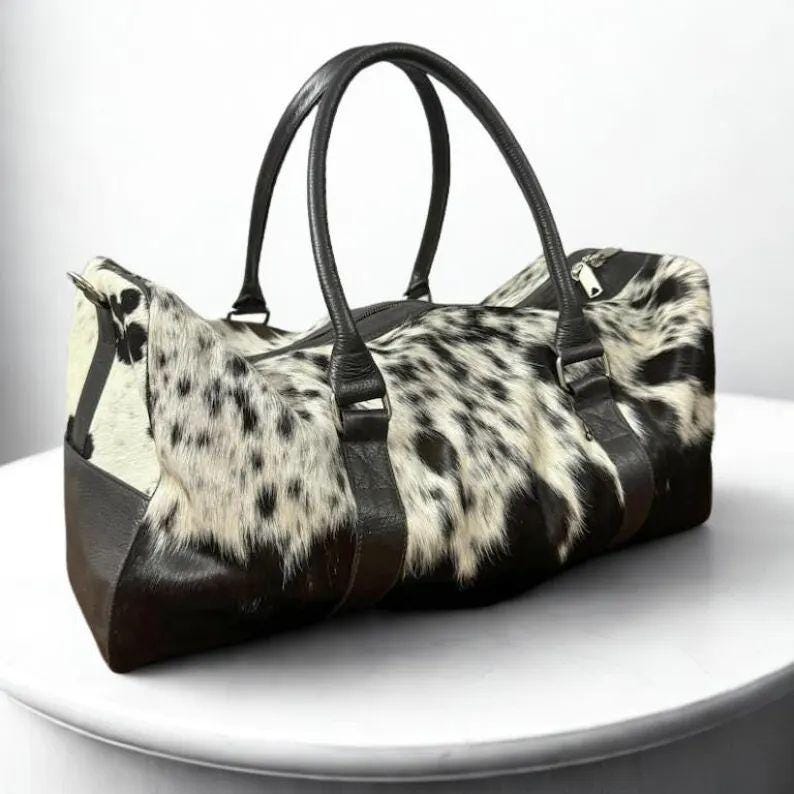 May include: A duffel bag with a spotted, black and white animal hide pattern. The bag has black leather handles and trim. The bag is designed with a zipper closure.