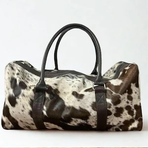 May include: A duffel bag with a cowhide pattern in shades of brown, black, and white. The bag has two black leather handles and a black zipper closure. The bag is rectangular and appears to be made of leather.