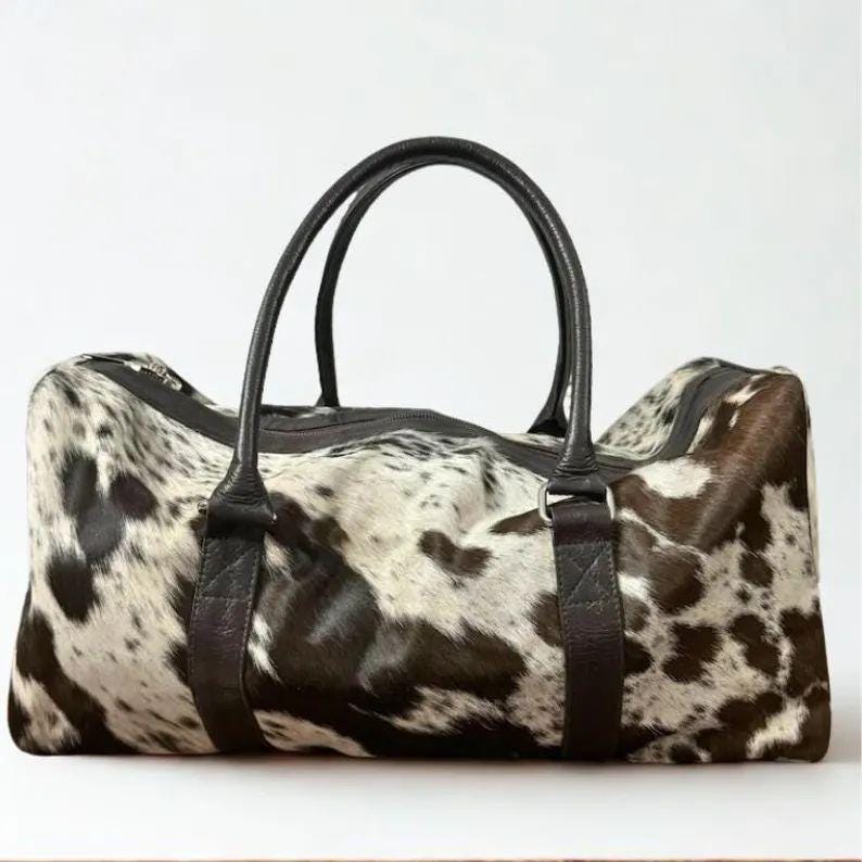 May include: A duffel bag with a cowhide pattern in shades of brown, black, and white. The bag has two black leather handles and a black zipper closure. The bag is rectangular and appears to be made of leather.