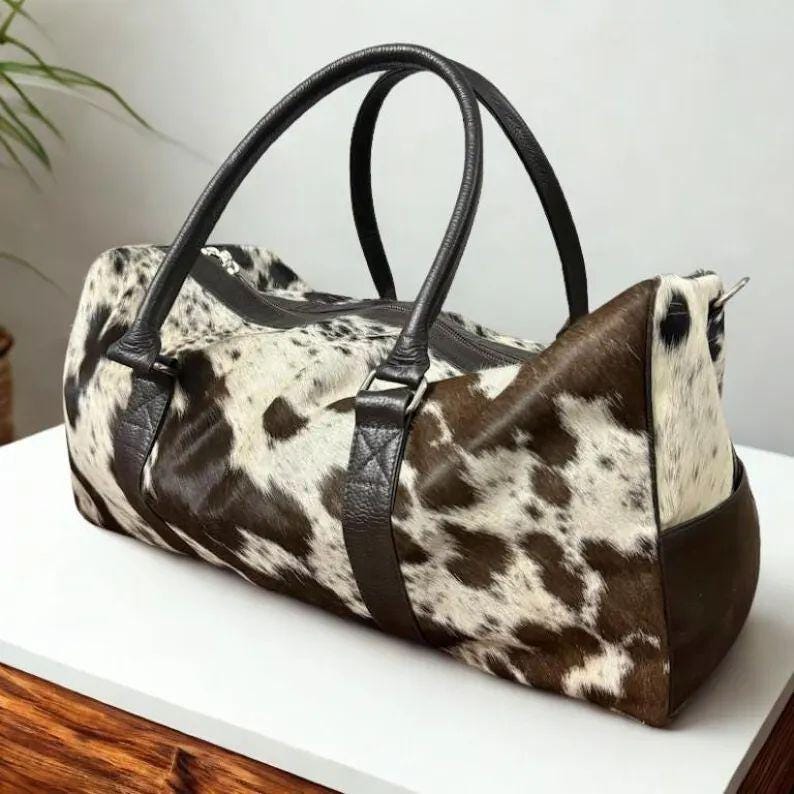 May include: A duffel bag with a cowhide pattern in shades of brown and white. The bag features black leather handles and trim, a zipper closure, and a side pocket. The bag is designed for travel or carrying essentials.