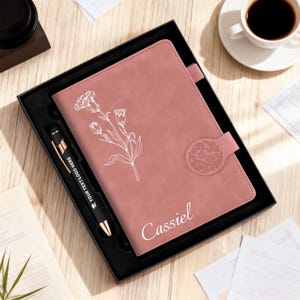 May include: A dusky pink journal and pen set in a black gift box. The journal has a floral design and the name "Cassiel". The black pen has gold accents and the text "YOUR TEXT LOGO HERE". A coffee cup is in the background.