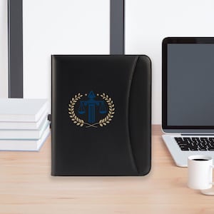 May include: Black leather portfolio with a gold and blue scales of justice emblem. The emblem is surrounded by a gold laurel wreath. The portfolio is on a wooden desk next to a laptop and a white mug.