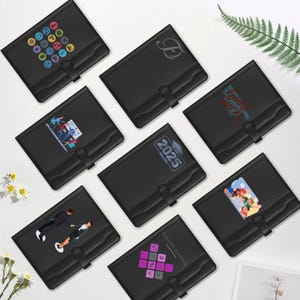 May include: A collection of black document holders with various designs. Some feature colorful graphics, a monogram, the year 2025, and the text "Sound Luck." Each holder has a strap closure and a pocket.