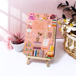 以下が含まれることがあります： ピンク色の長方形の装飾サイン。本をテーマにしたイラストと「Just one more chapter」の文字が描かれています。小さな木製のイーゼルに立てかけられています。背景は淡いピンク色です。