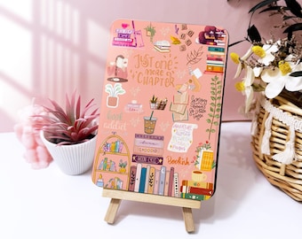 May include: A pink rectangular decorative sign with the text "Just one more chapter" and other book-related illustrations. The sign is propped up on a small wooden easel. The background is a soft pink.