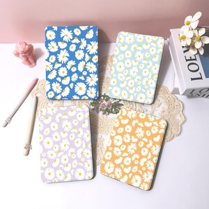 Puede incluir: Cuatro cuadernos rectangulares con estampados de margaritas. Los cuadernos tienen diferentes colores de fondo: azul, celeste, lavanda y naranja. Cada cuaderno presenta margaritas blancas con centros amarillos.
