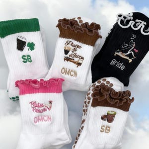 Könnte beinhalten: Eine Auswahl weißer Socken mit verschiedenen gestickten Designs und Texten. Eine Socke hat einen grünen Bund mit Bier und Kleeblatt, eine andere einen braunen Bund mit Kaffee und dem Text "Parted & Latte" und eine schwarze Socke mit dem Wort "Bride".