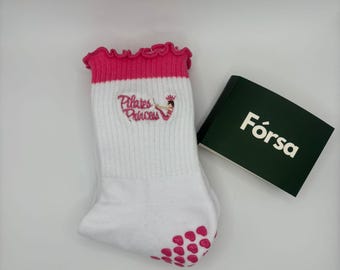 May include: White Pilates Princess socks with a pink ruffled top and pink non-slip grips on the sole. The socks have the words "Pilates Princess" embroidered on them. A green box with the word "Fórsa" is next to the socks.