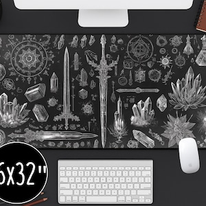 May include: Black desk mat featuring white illustrations of crystals, swords, and mystical symbols. The mat measures 40.6 x 81.3 cm. A white keyboard and mouse are visible.