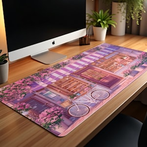 Puede incluir: Alfombrilla de escritorio rectangular con una ilustración detallada de una escena de calle europea. El diseño incluye una bicicleta, escaparates y flores rosas, con una paleta de colores de rosas, morados y marrones. La alfombrilla está colocada sobre un escritorio de madera.
