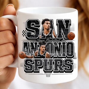 May include: White ceramic mug featuring the words "SAN ANTONIO SPURS" in a black and white design. The mug includes images of two basketball players and basketballs. The words "Spurs Basketball" are also printed on the mug.