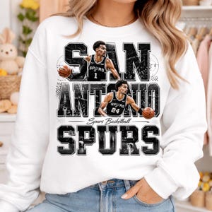 Pode incluir: Camisola branca com um design gráfico a preto e branco. O design apresenta as palavras "SAN ANTONIO SPURS", imagens de jogadores de basquetebol e o texto "Spurs Basketball".