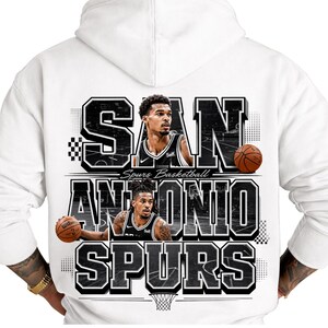 May include: White hoodie featuring a graphic design for the San Antonio Spurs basketball team. The design includes the team name, player images, and basketballs. The text "SAN ANTONIO SPURS" is prominently displayed.