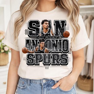 May include: Cream-colored t-shirt featuring a graphic design for the San Antonio Spurs basketball team. The design includes the team name, player images, and basketballs in black and white.