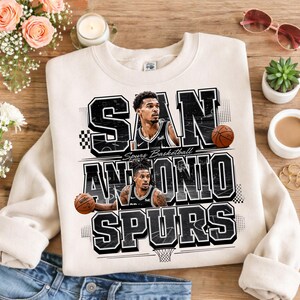 May include: Cream-colored San Antonio Spurs basketball sweatshirt. The design features the team name and two basketball players, along with basketballs. The text is in black and white, with a basketball hoop graphic at the bottom.
