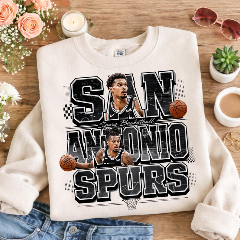 May include: Cream-colored San Antonio Spurs basketball sweatshirt. The design features the team name and two basketball players, along with basketballs. The text is in black and white, with a basketball hoop graphic at the bottom.