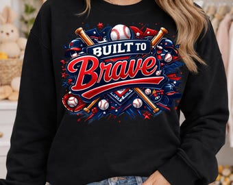 May include: Black sweatshirt featuring a baseball-themed graphic. The design includes baseballs, bats, and the text "BUILT TO Brave" in red and white. The graphic is set against a backdrop of blue and red stars and stripes.