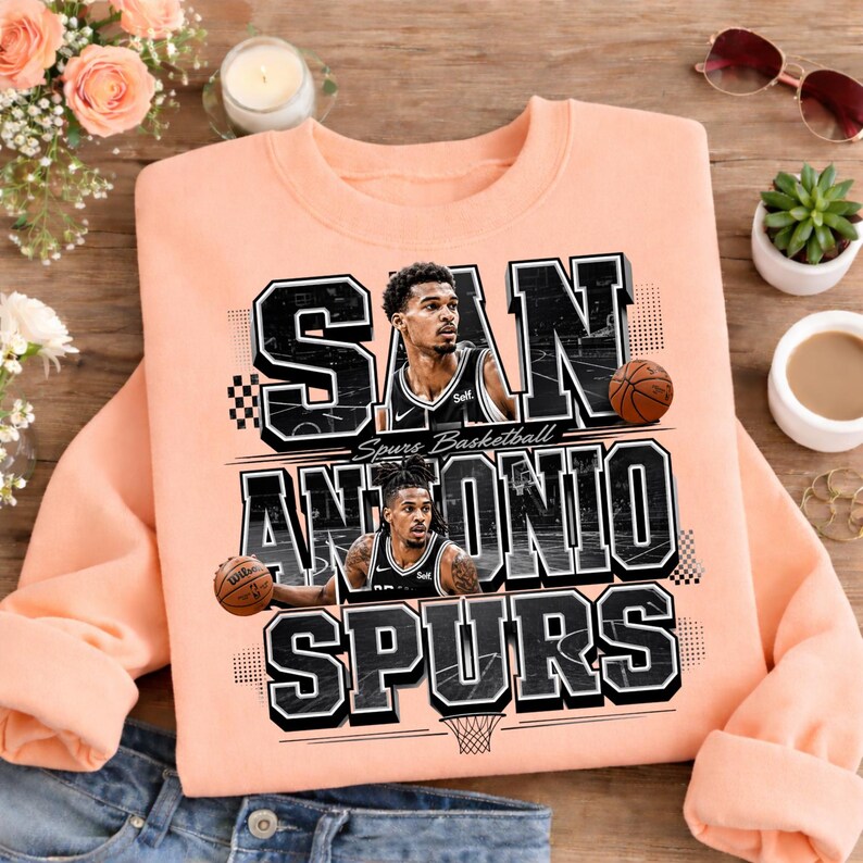 May include: A peach-colored sweatshirt featuring the text "SAN ANTONIO SPURS" in a black and white design. The sweatshirt includes images of two basketball players and a basketball. The words "Spurs Basketball" are also present.