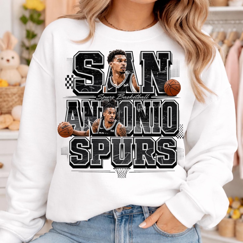 May include: White sweatshirt featuring the words "SAN ANTONIO SPURS" in a bold, black and white design. The graphic includes images of basketball players and basketballs. The words "Spurs Basketball" are also printed on the sweatshirt.