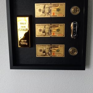 May include: A framed display featuring a gold bar, three gold-coloured £100 bills, two Bitcoin coins, and a car key. The gold bar is labelled "999.9 FINE GOLD" and weighs 1000g. The frame is black.