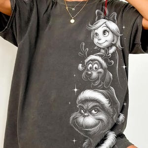 May include: Dark grey t-shirt featuring a black and white illustration of three Christmas characters stacked vertically. The top character is a girl with a bow, followed by a dog with antlers, and the Grinch at the bottom. The shirt has short sleeves.
