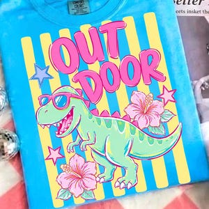 以下が含まれることがあります： サングラスをかけた緑色の恐竜のグラフィック、ピンク色の「OUT DOOR」の文字、ピンク色のハイビスカスの花が描かれた、ターコイズ色のComfort Colors Tシャツ。シャツには黄色と青の縦縞模様があります。