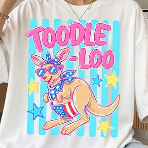 以下が含まれることがあります： サングラスとバンダナをつけた漫画のカンガルーが描かれた白いTシャツ。ピンク色の文字で「TOODLE-LOO」と書かれています。カンガルーは赤、白、青のデザインのポーチを持っており、青と黄色の星に囲まれています。