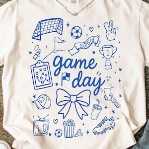 Puede incluir: Camiseta de color crema con un dise&ntilde;o gr&aacute;fico azul que presenta elementos relacionados con el f&uacute;tbol. El dise&ntilde;o incluye una red de f&uacute;tbol, un bal&oacute;n, un trofeo y las palabras "Game Day". Otras ilustraciones incluyen un silbato, palomitas de ma&iacute;z y una bota de f&uacute;tbol.