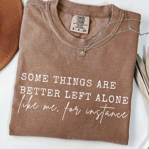 Puede incluir: Una camiseta marrón Comfort Colors con el texto "SOME THINGS ARE BETTER LEFT ALONE like me, for instance" en blanco. Un collar plateado con un pequeño colgante está sobre la camiseta.