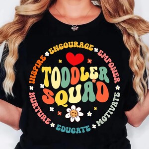 May include: Black t-shirt with a colorful graphic that says "Toddler Squad" in a retro font. The design includes a red heart, flowers, and the words "Inspire, Encourage, Empower, Motivate, Educate, Nurture".