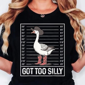 Może przedstawiać: Czarny t-shirt z kresk&oacute;wkową gęsią w kapeluszu i butach kowbojskich, na tle wykresu wzrostu. Gęś ma białe pi&oacute;ra i szare skrzydła. Tekst "GOT TOO SILLY" jest wyświetlany poniżej.