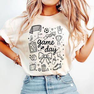 Puede incluir: Camiseta de color crema con un dise&ntilde;o gr&aacute;fico negro que presenta ilustraciones con tem&aacute;tica de f&uacute;tbol. El dise&ntilde;o incluye una porter&iacute;a de f&uacute;tbol, un bal&oacute;n, un trofeo y las palabras "game day". Otros elementos incluyen un lazo, palomitas de ma&iacute;z y una bota de f&uacute;tbol.