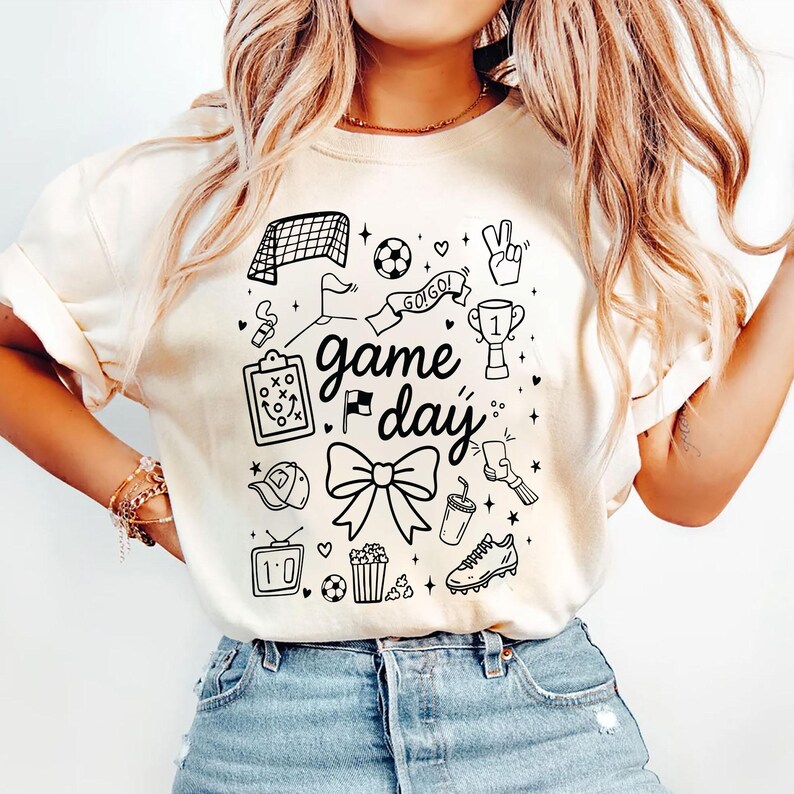Puede incluir: Camiseta de color crema con un dise&ntilde;o gr&aacute;fico negro que presenta ilustraciones con tem&aacute;tica de f&uacute;tbol. El dise&ntilde;o incluye una porter&iacute;a de f&uacute;tbol, un bal&oacute;n, un trofeo y las palabras "game day". Otros elementos incluyen un lazo, palomitas de ma&iacute;z y una bota de f&uacute;tbol.