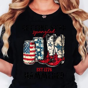 May include: Black t-shirt with a patriotic design featuring two beer cans and a pair of cowboy boots. The cans are decorated with the American flag and stars. The boots are white with blue stars and red accents. The text reads "Getting Star Spangled, EST. 1776, Hammered."