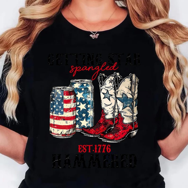 May include: Black t-shirt with a patriotic design featuring two beer cans and a pair of cowboy boots. The cans are decorated with the American flag and stars. The boots are white with blue stars and red accents. The text reads "Getting Star Spangled, EST. 1776, Hammered."