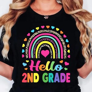 May include: A black t-shirt featuring a colorful graphic of a rainbow with leopard print and polka dot stripes. A pink heart sits in the center of the rainbow, surrounded by small hearts. The text "Hello 2ND GRADE" is printed in bright, multi-colored letters.