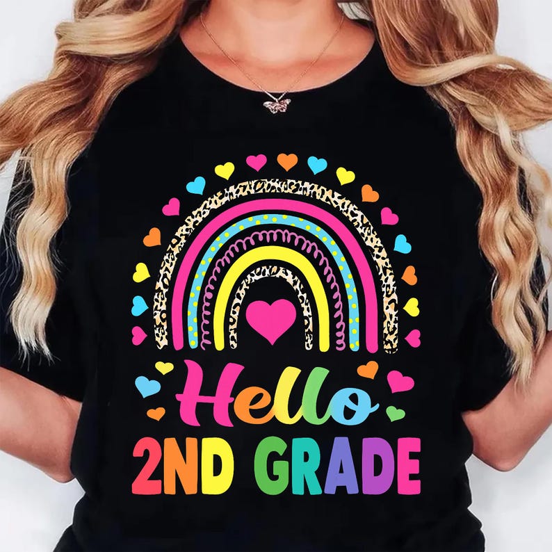 May include: A black t-shirt featuring a colorful graphic of a rainbow with leopard print and polka dot stripes. A pink heart sits in the center of the rainbow, surrounded by small hearts. The text "Hello 2ND GRADE" is printed in bright, multi-colored letters.