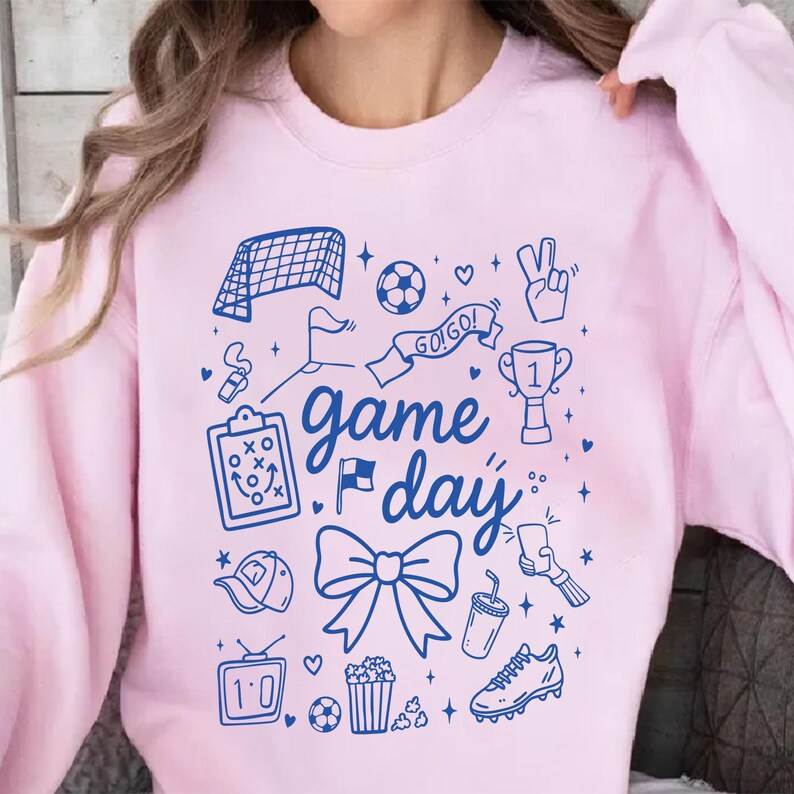 Puede incluir: Sudadera rosa claro con un dise&ntilde;o gr&aacute;fico azul. El dise&ntilde;o incluye ilustraciones tem&aacute;ticas de f&uacute;tbol como una porter&iacute;a, un bal&oacute;n de f&uacute;tbol, un trofeo y las palabras "game day". Otros elementos incluyen un lazo, palomitas de ma&iacute;z y un televisor.
