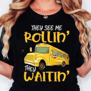 May include: Black t-shirt featuring a yellow school bus graphic with the text "They See Me Rollin', They Waitin'". The bus has a "STOP" sign. The text is in a playful, handwritten style. The shirt is a crew neck.