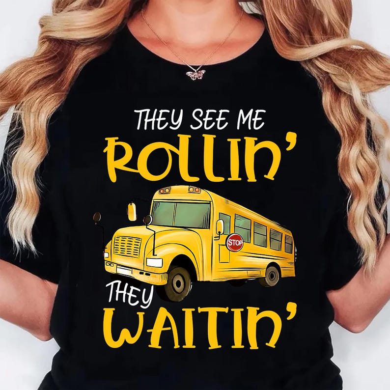 May include: Black t-shirt featuring a yellow school bus graphic with the text "They See Me Rollin', They Waitin'". The bus has a "STOP" sign. The text is in a playful, handwritten style. The shirt is a crew neck.