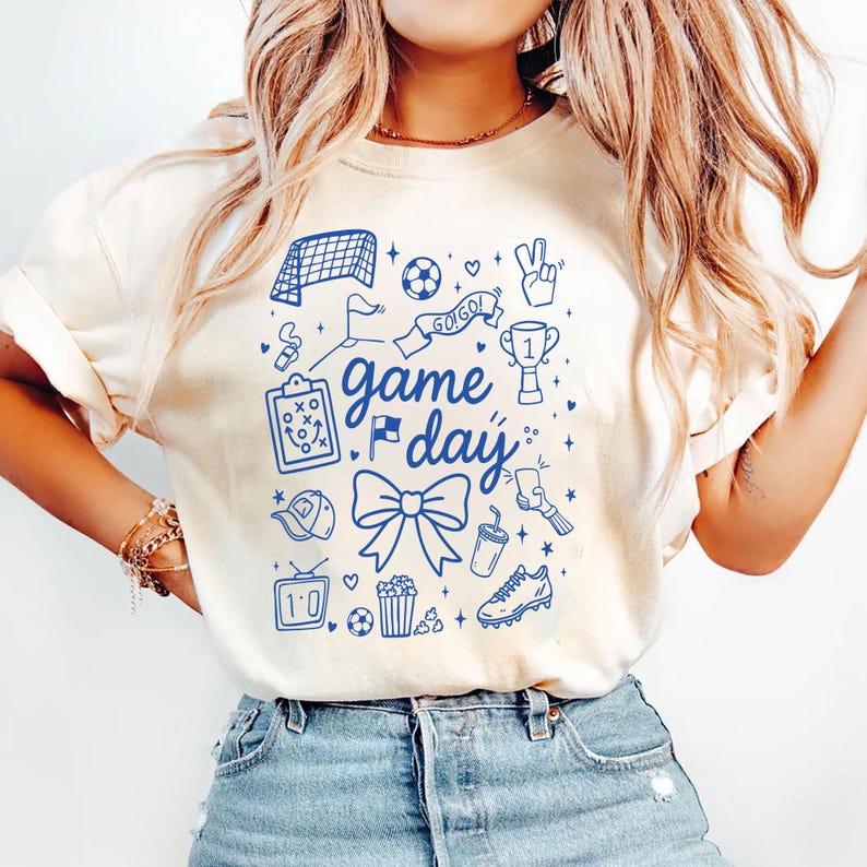 Puede incluir: Camiseta color crema con un dise&ntilde;o gr&aacute;fico azul que presenta iconos relacionados con el f&uacute;tbol y las palabras "Game Day". El dise&ntilde;o incluye una red de f&uacute;tbol, un bal&oacute;n, un trofeo y un lazo. La camiseta es de material suave.