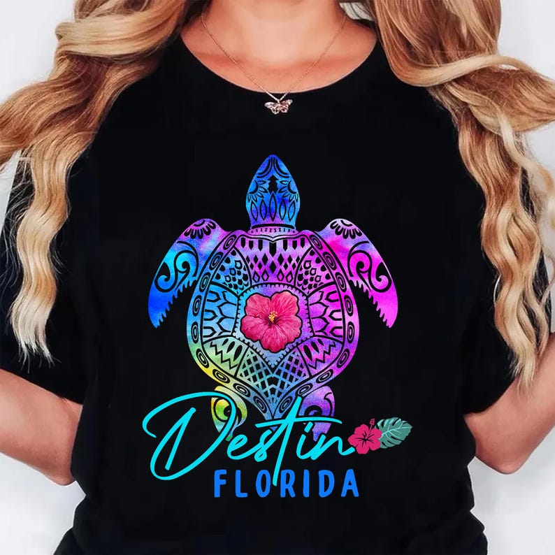 May include: Black t-shirt featuring a colorful sea turtle design with a hibiscus flower at its center. The turtle is in shades of blue, purple, and pink. The text "Destin FLORIDA" is written in a turquoise script font.