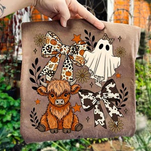 Op de afbeelding: Een bruin kussen met een cartoon Highland koe, een spook en twee strikken met pompoen- en koeienpatronen. Het ontwerp bevat oranje sterren, bloemen en bladeren op een bruine achtergrond.
