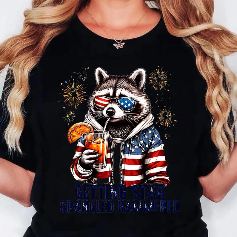 May include: Black t-shirt featuring a raccoon wearing sunglasses with American flag designs. The raccoon is holding a drink with an orange slice. The shirt has the text "GETTING STAR SPANGLED HAMMERED" in blue.